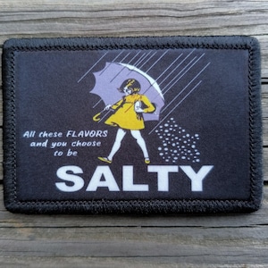 May include: Black rectangular patch with a black border. The patch features a cartoon illustration of a person in a yellow dress holding a purple umbrella, with the word "SALTY" in white. Text reads "All these FLAVORS and you choose to be".