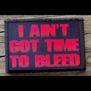May include: A rectangular patch with the text "I AIN'T GOT TIME TO BLEED" in red, block letters. The patch has a black background and a black border. The patch is on a wooden surface.
