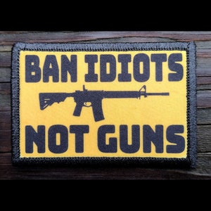 May include: A rectangular patch with a yellow background and black text that reads "BAN IDIOTS NOT GUNS." A black silhouette of a rifle is centered on the patch. The patch has a black border.
