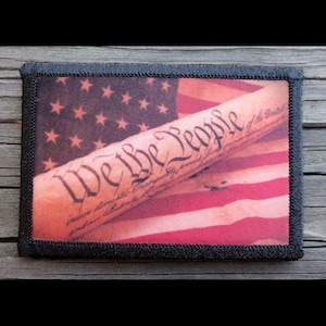 May include: A black and white patch with a red, white, and blue American flag in the background. The words "We the People" are printed on the patch in a stylized font.