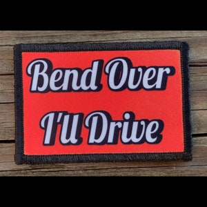 May include: Rectangular patch with the text "Bend Over I'll Drive" in white script on a red background. The patch has a black border and is displayed on a wooden surface.