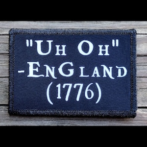 May include: A rectangular navy blue patch with white text. The text reads "Uh Oh" above "- England (1776)". The patch has a black stitched border and is set against a wooden background.