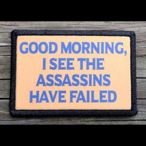 May include: Rectangular patch with the text "GOOD MORNING, I SEE THE ASSASSINS HAVE FAILED" in blue on a peach background. The patch has a black border and is set against a wooden surface.