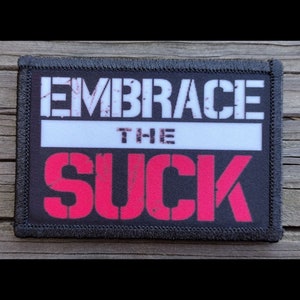 May include: Black rectangular patch with the words "EMBRACE THE SUCK" in white and red block letters. The patch has a black border and is set against a wooden background.