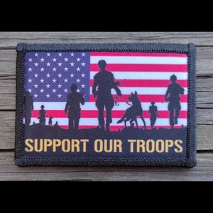 May include: A black and white embroidered patch with a silhouette of soldiers and a dog walking in front of an American flag. The text "SUPPORT OUR TROOPS" is printed in gold below the image.
