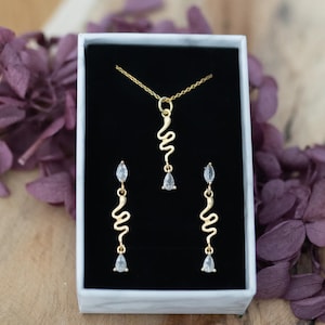 Könnte beinhalten: Ein goldenes Halsketten- und Ohrring-Set mit einem zarten, wellenförmigen Design und klaren Steinen. Der Schmuck ist in einer weißen Schachtel mit schwarzem Innenraum ausgestellt.