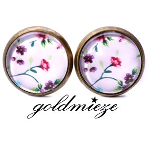 Könnte beinhalten: Ein Paar bronzefarbene Ohrringe mit einem floralen Design. Die Ohrringe haben einen runden, gläsernen Cabochon mit einem rosa und violetten Blumenmuster auf weißem Hintergrund.