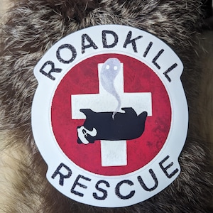 Könnte beinhalten: Ein runder Aufkleber mit den Worten "ROADKILL RESCUE" in Weiß. Das Design zeigt ein rotes Kreuz auf weißem Hintergrund, mit einem schwarz-weißen Dachs und einer Geisterillustration.