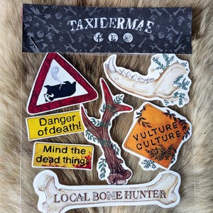 Könnte beinhalten: Eine Sammlung von Aufklebern mit verschiedenen Designs. Dazu gehören ein Straßenschild mit einem Dachs, ein Knochen, ein Kieferknochen und Texte wie "Danger of death!", "Mind the dead things", "Vulture Culture" und "Local Bone Hunter".
