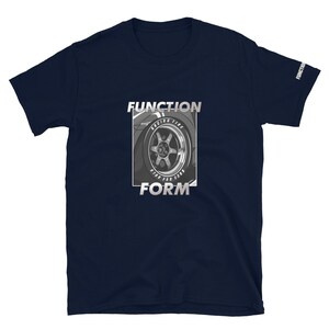 Op de afbeelding: Marineblauw T-shirt met korte mouwen met een afbeelding van een autowiel en de woorden "FUNCTION" en "FORM" in zilver. De mouw heeft het woord "FUNCTION" erop gedrukt. Het ontwerp staat in een rechthoekige lijst.
