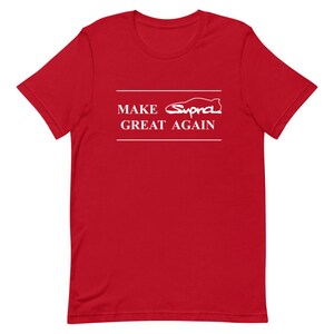 May include: A red t-shirt featuring white text and a car graphic. The text says "MAKE Supra GREAT AGAIN", with "Supra" represented by a car silhouette. The shirt is a short-sleeved crew neck.