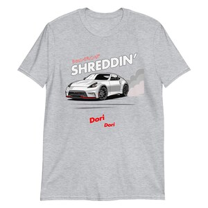 May include: A light grey t-shirt with a white sports car graphic and the text "SHREDDIN'" above it, with Japanese characters. Below the car, the text "Dori Dori" is in red. The car has red detailing.