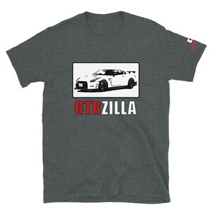 Könnte beinhalten: Dunkelgraues T-Shirt mit einer weißen Autoillustration in einem schwarzen Rechteck. Der Text "GTRZILLA" befindet sich unter dem Auto in Rot und Weiß. Eine kleine japanische Flagge befindet sich auf dem Ärmel.