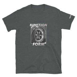 Op de afbeelding: Donkergrijs t-shirt met een afbeelding van een autowiel en de woorden "FUNCTION" en "FORM" in een rechthoekige lijst. De wielafbeelding is in grijstinten en wit, met de woorden "RACING TIRE" en andere tekst zichtbaar.