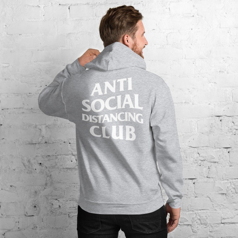Peut inclure: Un sweat &agrave; capuche gris clair avec le texte blanc "ANTI SOCIAL DISTANCING CLUB" imprim&eacute; au dos. Le sweat &agrave; capuche a une capuche et des manches longues. La personne sur l'image porte un pantalon noir.