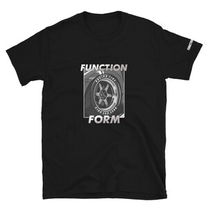 Op de afbeelding: Zwart T-shirt met korte mouwen met een afbeelding van een autowiel en de woorden "FUNCTION" en "FORM" in een zilverkleurig, omlijnd lettertype. De woorden "RACING TIRE" zijn ook zichtbaar. Het woord "FUNCTION" staat op de mouw.