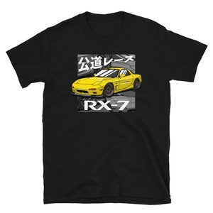 May include: Black t-shirt with a yellow sports car graphic. The graphic includes Japanese text and the text "RX-7". The car is illustrated in a cartoon style, set against a grey background.