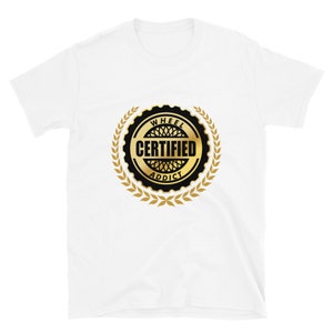 May include: White t-shirt featuring a gold and black circular design with the words "WHEEL CERTIFIED ADDICT" in a gold and black emblem. The design is surrounded by a gold laurel wreath.