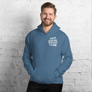 Peut inclure: Un sweat &agrave; capuche bleu avec une poche avant et une capuche. Le texte "ANTI SOCIAL DISTANCING CLUB" est imprim&eacute; en blanc sur la poitrine. La personne sur la photo sourit.