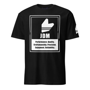 May include: Black t-shirt with a white graphic featuring a stylized design and the letters "JDM." Below the graphic, the text reads "Performance. Quality. Craftsmanship. Precision. Enjoyment. Reliability."