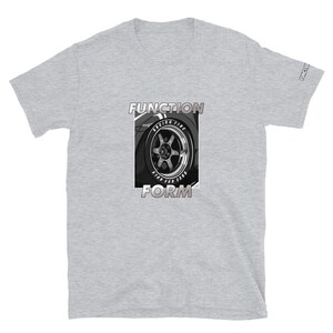 Op de afbeelding: Lichtgrijs t-shirt met een afbeelding van een autowiel en de woorden "FUNCTION" en "FORM" in een vetgedrukte lettertype. De wielafbeelding is in zwart en wit, met "RACING TIRE" zichtbaar. De mouw heeft het woord "FUNCTION" erop gedrukt.