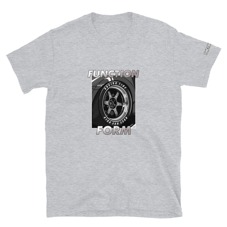 Op de afbeelding: Lichtgrijs t-shirt met een afbeelding van een autowiel en de woorden "FUNCTION" en "FORM" in een vetgedrukte lettertype. De wielafbeelding is in zwart en wit, met "RACING TIRE" zichtbaar. De mouw heeft het woord "FUNCTION" erop gedrukt.