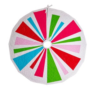 Pode incluir: Uma saia de árvore de Natal acolchoada branca com um design de raios de sol coloridos. A saia apresenta listras alternadas de rosa, vermelho, verde e azul.