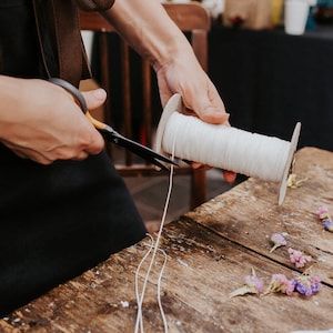 May include: A person is cutting white string from a spool using a pair of scissors with yellow handles. The string is lying on a wooden table with dried flowers scattered around.