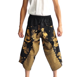 May include: Black and gold patterned cropped pants with a drawstring waist. The pants feature a repeating gold koi fish and floral design. The model is wearing a white tank top.