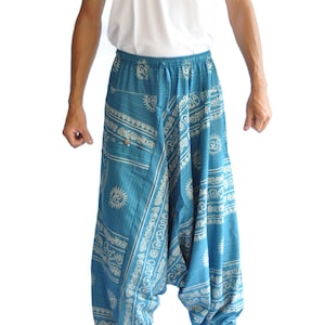 Op de afbeelding: Tealblauwe harembroek met een wit printpatroon van zonnen en Om-symbolen. Deze ruimvallende broek heeft een elastische tailleband met trekkoord, een laaghangend kruis, elastische enkelboorden en een kleine zak met een knoop aan de voorzijde.