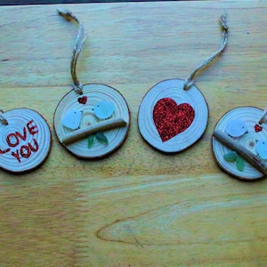 Könnte beinhalten: Holzornamente mit Kordelaufhängern. Ein Ornament trägt die Worte "LOVE YOU" in rotem Glitzer. Andere Ornamente zeigen weiße Vögel auf einem Ast, ein rotes Glitzerherz und kleine grüne Blätter. Perfekt für den Valentinstag.