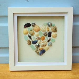 May include: A white framed picture with a heart shape made of seashells and sea glass. The heart is on a light green background.