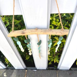 May include: A driftwood mobile with strands of sea glass in shades of green and white. The sea glass is suspended from a natural wood branch with twine. The mobile hangs against a white picket fence with green foliage in the background.