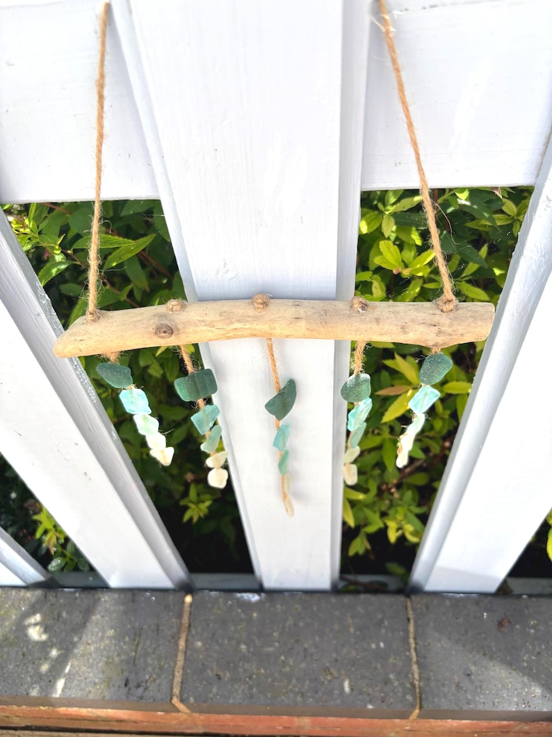 May include: A driftwood mobile with strands of sea glass in shades of green and white. The sea glass is suspended from a natural wood branch with twine. The mobile hangs against a white picket fence with green foliage in the background.