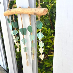 May include: A handcrafted wind chime featuring sea glass in shades of green and white, suspended from a natural driftwood branch with jute twine. The sea glass pieces are various shapes and sizes, creating a soothing, coastal aesthetic.