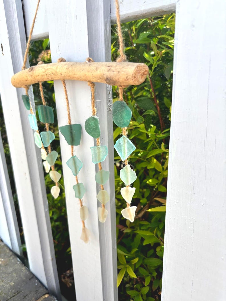 May include: A handcrafted wind chime featuring sea glass in shades of green and white, suspended from a natural driftwood branch with jute twine. The sea glass pieces are various shapes and sizes, creating a soothing, coastal aesthetic.