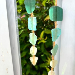 May include: A handcrafted wind chime featuring sea glass pieces in shades of green and white, strung on natural twine. The sea glass is suspended from a piece of driftwood, creating a coastal-themed decorative item.