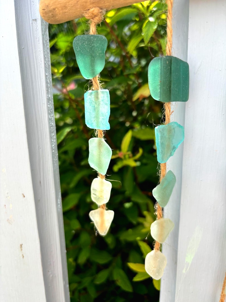 May include: A handcrafted wind chime featuring sea glass pieces in shades of green and white, strung on natural twine. The sea glass is suspended from a piece of driftwood, creating a coastal-themed decorative item.