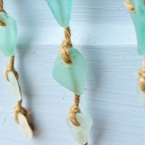 May include: Close-up of a decorative hanging strand featuring sea glass in shades of white and aqua. The sea glass pieces are strung on natural twine, creating a coastal or beach-themed aesthetic. The background is a light blue painted surface.