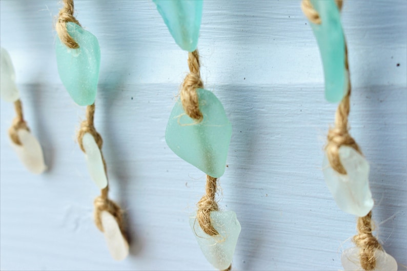 May include: Close-up of a decorative hanging strand featuring sea glass in shades of white and aqua. The sea glass pieces are strung on natural twine, creating a coastal or beach-themed aesthetic. The background is a light blue painted surface.