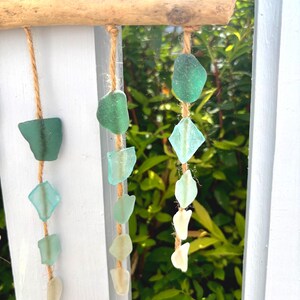 May include: A decorative hanging mobile featuring sea glass in shades of green and white, suspended from a natural wooden branch with twine. The sea glass pieces vary in shape and size, creating a coastal aesthetic.