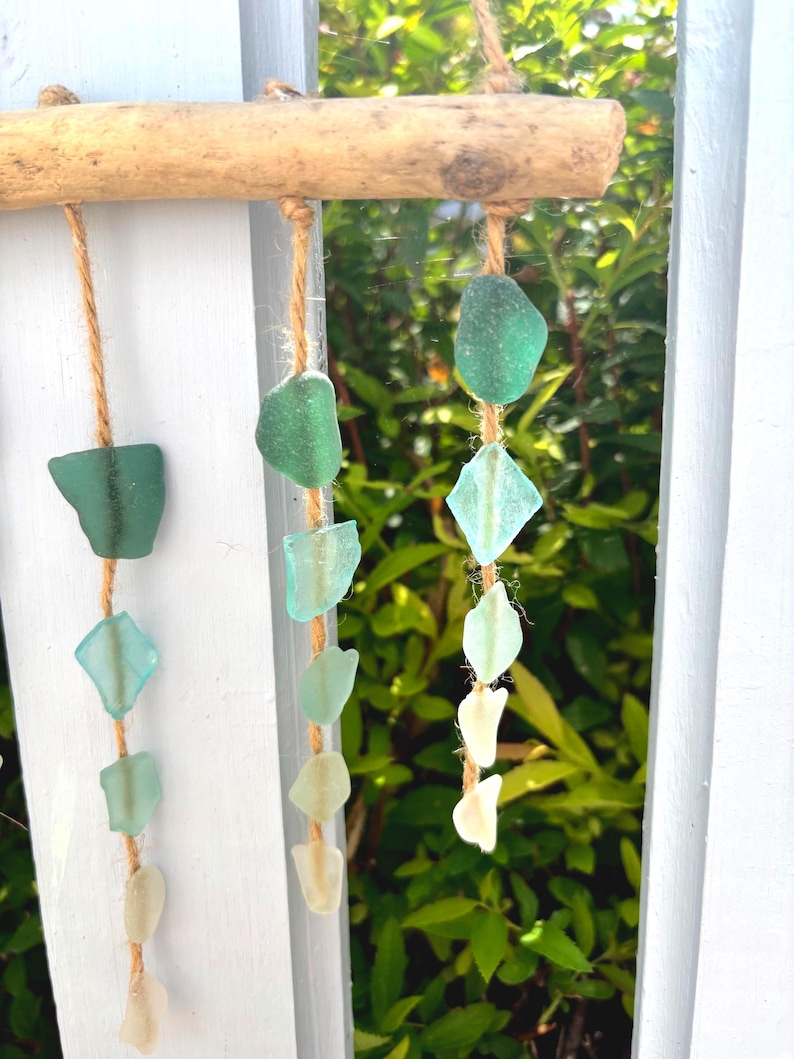 May include: A decorative hanging mobile featuring sea glass in shades of green and white, suspended from a natural wooden branch with twine. The sea glass pieces vary in shape and size, creating a coastal aesthetic.