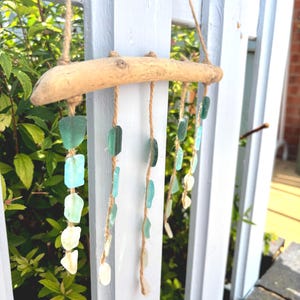 May include: A handcrafted wind chime made from a piece of driftwood and strands of sea glass. The sea glass pieces are in shades of green, blue, and white, and are suspended from the driftwood with twine. The wind chime hangs on a white picket fence.