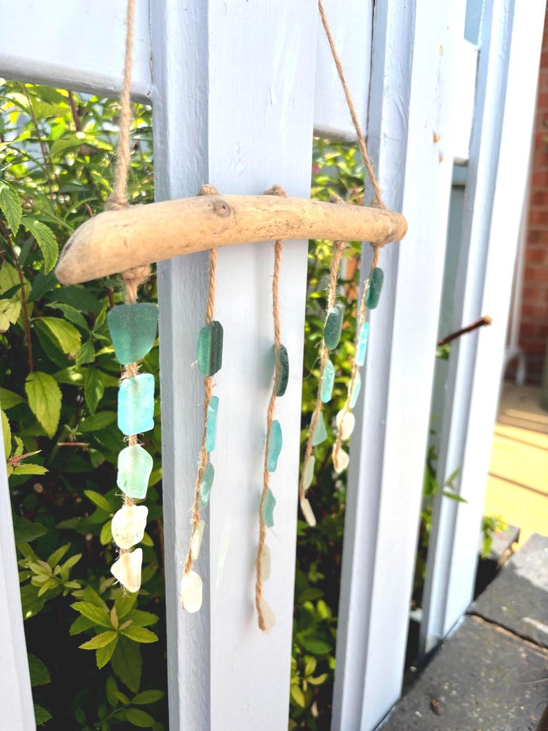 May include: A handcrafted wind chime made from a piece of driftwood and strands of sea glass. The sea glass pieces are in shades of green, blue, and white, and are suspended from the driftwood with twine. The wind chime hangs on a white picket fence.