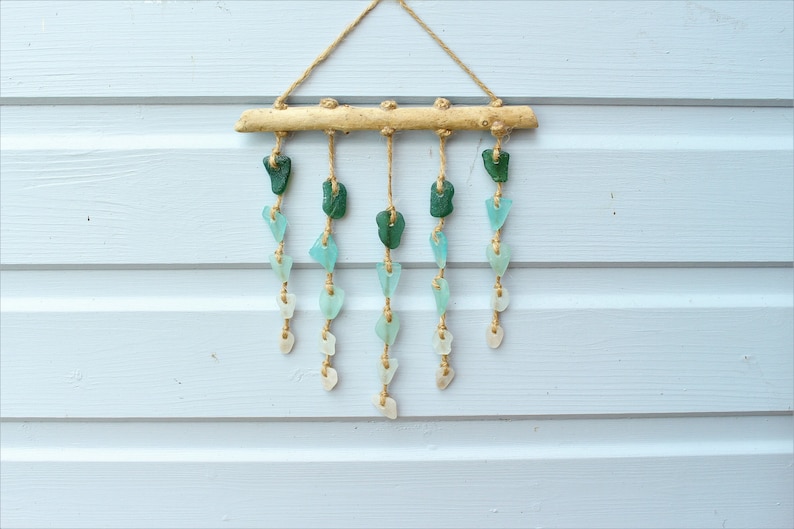 May include: A decorative wall hanging featuring sea glass in shades of green, blue, and white. The sea glass pieces are strung on twine and suspended from a natural wood branch. The background is a light blue wooden wall.