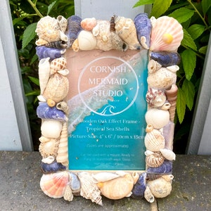 May include: A wooden picture frame decorated with seashells. The frame is 4 inches by 6 inches and has a light blue background with a wave design. The text "CORNISH MERMAID STUDIO Coastal Decor" is printed on the frame.