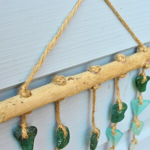 May include: A rustic wall hanging featuring a natural wooden branch with jute twine and sea glass accents. The sea glass pieces are in shades of green and blue, adding a coastal touch. The piece is suspended by jute rope.
