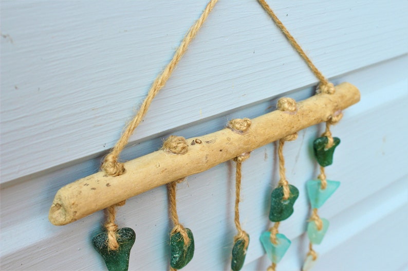 May include: A rustic wall hanging featuring a natural wooden branch with jute twine and sea glass accents. The sea glass pieces are in shades of green and blue, adding a coastal touch. The piece is suspended by jute rope.