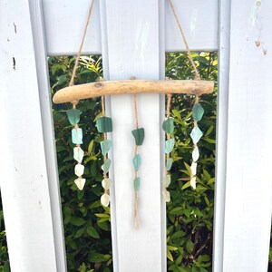 May include: A driftwood wind chime with strands of sea glass in shades of green and white. The sea glass pieces are strung on natural twine and suspended from a piece of driftwood. The wind chime hangs against a white picket fence.