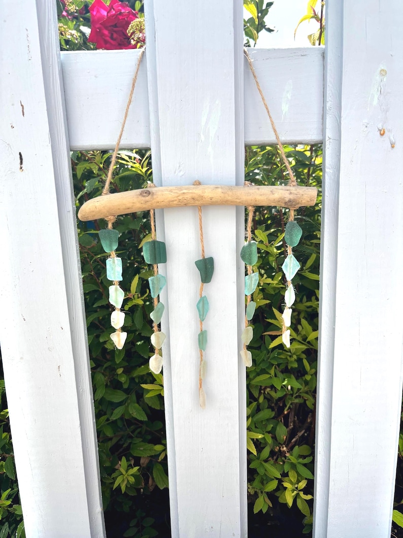 May include: A driftwood wind chime with strands of sea glass in shades of green and white. The sea glass pieces are strung on natural twine and suspended from a piece of driftwood. The wind chime hangs against a white picket fence.